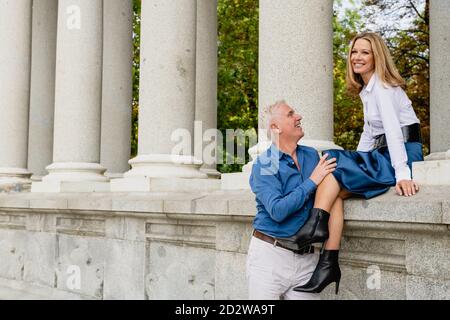 Entzückter Mann, der in der Nähe der Steingrenze steht und die Beine einer Frau, die während eines Spaziergangs im Park auf dem Zaun sitzt, zärtlich berührt Stockfoto