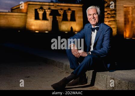 Positiv eleganter Senior-Rüde im stilvollen blauen Anzug mit Schleife Krawatte und Armbanduhren sitzen auf Steinrand in der Stadt der Nacht Und Blick auf die Kamera Stockfoto
