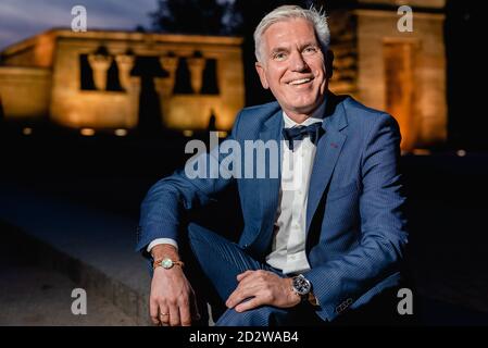 Positiv eleganter Senior-Rüde im stilvollen blauen Anzug mit Schleife Krawatte und Armbanduhren sitzen auf Steinrand in der Stadt der Nacht Und Blick auf die Kamera Stockfoto