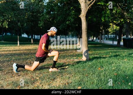 Seitenansicht des konzentrierten männlichen Athleten in Sportbekleidung beim Ausfallschritt Und Strecken der Beine beim Aufwärmen während des Trainings im Park Stockfoto