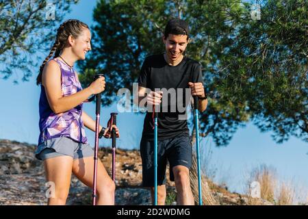 Niedriger Winkel von glücklichen jungen Mann und Frau in aktiver Wear mit Trekkingstöcken, die beim Stehen auf Berghang beim Wandern Pause machen und gemeinsam am Sommertag in der Natur laufen Stockfoto