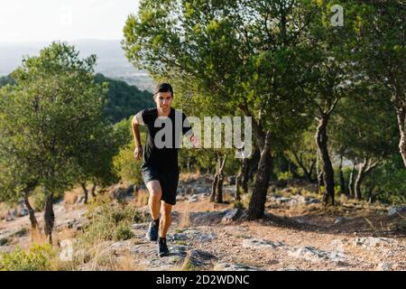 Positiver junger Athletic Mann im activewear Laufen auf steinigen Trail In grün bewaldeten bergigen Gelände Stockfoto