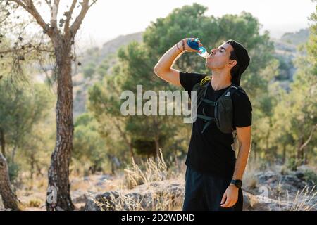 Junger männlicher Wanderer in aktivem Trinkwasser aus Plastikflasche Beim Ausruhen während des Trailrunning auf bergigem Gelände mit Grün Wald Stockfoto