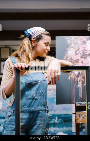 Kreative junge Künstlerin in Denim-Overall lehnt sich an leer Rahmen in Werkstatt mit Gemälden Stockfoto