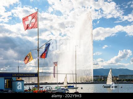 Die Schweizer Flagge fliegt an einem stürmischen Tag am Ufer des Genfersees in Genf, Schweiz, mit dem Jet d'Eau Wasserstrahlbrunnen in der Ferne Stockfoto