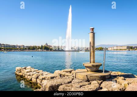 Die Stadt und die Bucht von Genf, Schweiz, mit ihrem Wasserstrahlbrunnen auf dem Genfer See, von einem Steg mit einem Leuchtfeuer an einem sonnigen Sommermorgen gesehen. Stockfoto