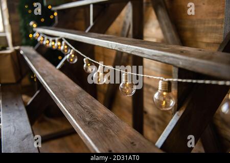 Weihnachtsgirlande von Glühbirnen, die von einer Holztreppe hängen. Festliche Heimtextilien. Skandinavischer Loft-Stil.Textur Hintergrund, Kopierraum Stockfoto