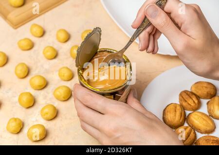 Frau Hand nimmt einen Teelöffel gekochte Kondensmilch aus einer Dose, neben rohem Plätzchenteig und fertigen Hälften für Plätzchen Nüsse Stockfoto