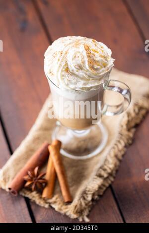 Kürbis Gewürz Latte in einem Glas Becher. Kürbislatte mit Schlagsahne und Gewürzen auf Holzgrund. Heißer Herbstkaffee auf einer Sackleine Stockfoto