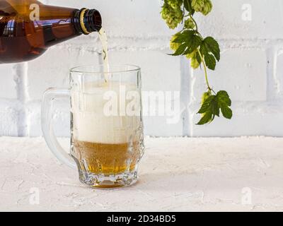 Der Glaskrug wird mit Bier aus einer Plastikflasche gefüllt. Ein Hopfenzweig und eine weiße Backsteinmauer auf dem Hintergrund. Stockfoto