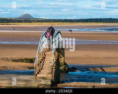 Belhaven, East Lothian, Schottland, Großbritannien, 7. Oktober 2020. UK Wetter: Sonnenschein auf der Bridge to Nowhere. Die Brücke über das Bieler Wasser ist nur bei Ebbe für Besucher des Strandes an der Belhaven Bay mit Blick auf Berwick Law in der Ferne zugänglich. Ein Paar geht über die ungewöhnliche Brücke Stockfoto