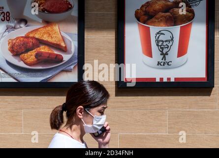 Eine Frau mit Gesichtsmask spricht auf ihrem Smartphone, während sie an der amerikanischen Fast-Food-Hühnchen-Restaurantkette Kentucky Fried Chicken (KFC) und dem Logo in Hongkong vorbeigeht. Stockfoto