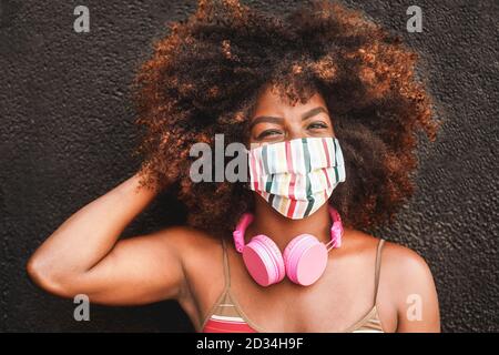Glückliche afrikanische Frau mit Kopfhörern trägt Gesichtsmaske während Coronavirus Outbreak - Fokus auf das Gesicht des Mädchens Stockfoto
