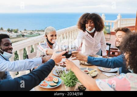 Junge, multirassische Menschen jubeln mit Wein, während sie Schutzmasken tragen - Glückliche Freunde, die Spaß beim Abendessen auf der Terrasse des Restaurants haben Outdoor - Socia Stockfoto