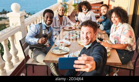Junge multirassische Menschen, die Selfie machen, während sie Schutzmasken tragen - Glückliche Freunde, die Spaß beim Abendessen auf der Terrasse im Freien haben - soziale Distanz Stockfoto