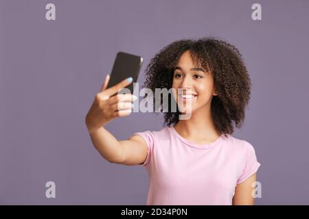 Moderne Technologie, mobile App und Selfie für Fashion Blog. Lächelnde afroamerikanische Frau macht Foto auf Smartphone isoliert auf lila Hintergrund Stockfoto