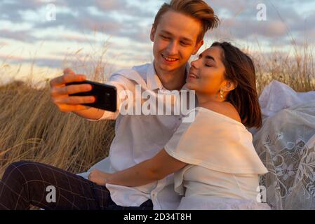 Selfie von Ehemann und Ehefrau von jungen schönen Paar von zweiundzwanzig Jahren. Porträt eines Mannes mit einem Mädchen. Sitzen im Gras mit einem Smartphone macht Stockfoto