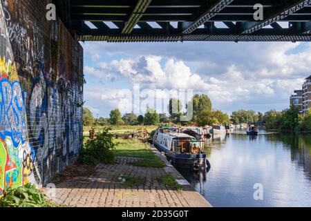 Großbritannien, London. Stadtlandschaft - Hausboote auf dem Fluss Lea schiffbaren Kanalabschnitt mit Graffiti unter einer Eisenbahnbrücke und einem klaren Sommerhimmel Stockfoto