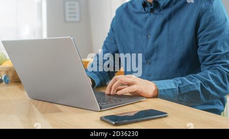 Nahaufnahme eines Mannes, der zu Hause am Tisch sitzt und an seinem modernen Silver Laptop arbeitet. Smartphone liegt auf einem Holztisch neben ihm. Stockfoto