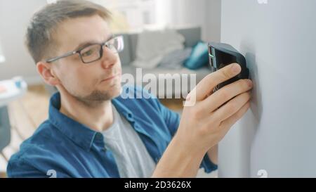 Ein junger Mann in Brille trägt ein blaues Hemd und hängt eine moderne Bewegungsmelder-Einheit an eine weiße Wand zu Hause. Gerät blinkt mit grüner LED-Anzeige. Stockfoto