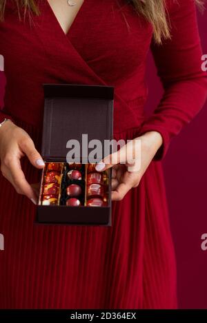 Nahaufnahme einer kleinen Schachtel Bonbons in weiblichen Händen. Frau, die Schokolade Süßigkeiten in kleine Box. Roter Studiohintergrund. Wohlschmeckende Geschenke für C Stockfoto