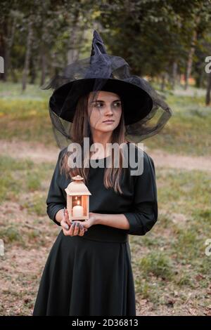 Junge Frau in dunklem Kleid und Hexenhut hält Laterne mit Kerzen in den Händen und beleuchtet den Wald. Halloween Party Kostüm. Parken mit Autum Stockfoto