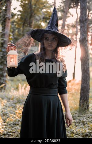 Junge Frau in dunklem Kleid und Hexenhut hält Laterne mit Kerzen in den Händen und beleuchtet den Wald. Halloween Party Kostüm. Parken mit Autum Stockfoto