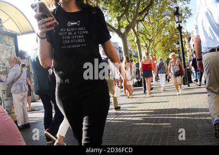 Junge Frau hält ein Handy, während sie in einer Straße der Stadt läuft. Go vegan schwarzes Shirt. Barcelona, Katalonien, Spanien. Stockfoto