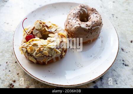 Süße Donuts auf einem Teller auf einem Marmortisch. Ein Teller mit zwei leckeren glasierten Donuts mit Schokolade, Nüssen und roter Kirsche. Stockfoto