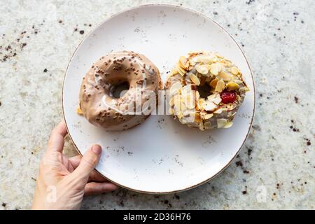 Süße Donuts auf einem Teller auf einem Marmortisch. Junge Frauenhand hält einen Teller mit zwei leckeren glasierten Donuts mit Schokolade, Nüssen und roter Kirsche Stockfoto