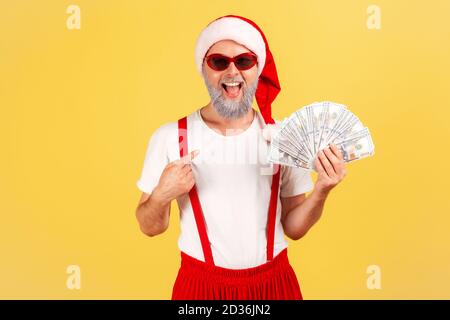 Extrem glücklich aufgeregt erwachsenen Mann in weihnachtsmann Hut und stilvolle Sonnenbrille zeigt Finger auf Dollar in der Hand, Vergünstigungen und Boni im Urlaub. Ind Stockfoto