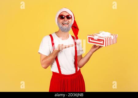 Extrem glücklich graubärtiger Mann in Sonnenbrille und weihnachtsmann Kostüm zeigt geschenkbox Blick auf die Kamera mit toothy Lächeln. Innenaufnahmen im Studio Isolate Stockfoto