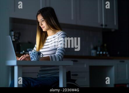 Junge Frau im Rollstuhl, die bis spät in die Nacht am Laptop arbeitet Stockfoto