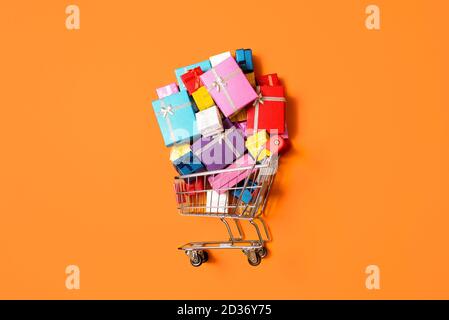 Warenkorb mit einem Stapel von bunten Geschenken auf orangefarbenem Hintergrund isoliert. Draufsicht mit Supermarkt-Wagen mit Geschenken. Weihnachtseinkaufskonzept. Stockfoto