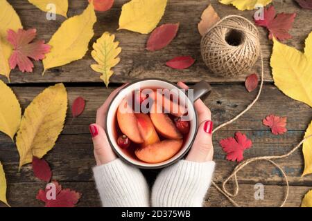 Damenhänden mit einem weißen Pullover halten Tassen mit einem warmen Fruchtgetränk auf einem hölzernen Hintergrund mit herbstlichen gelben und roten Blättern. Stockfoto