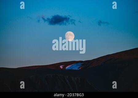 Atmosphärische Mondlandschaft der Dämmerung in Bergen. Dunkler Flieder Himmel und großer Mond mit Wolke über Silhouetten von Bergen in der Dämmerung. Schnee auf Felsen in m Stockfoto