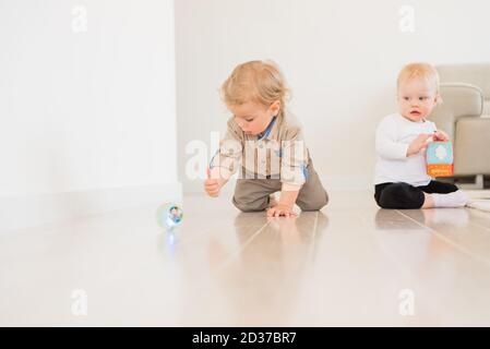 Niedliches Kleinkind Baby Junge kriechen auf dem Boden zu Hause und spielen mit bunten Ball. Baby Mädchen sitzen und spielen im Hintergrund. Stockfoto