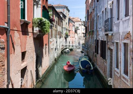 2015: Mann segelt an einem sonnigen Tag im roten Motorboot durch einen sehr engen Venedig-Kanal. Traditionelles Stadtbild Von Venedig Stockfoto