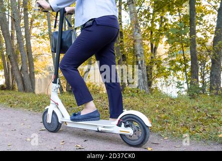 Eine Erwachsene elegante Frau ist im Begriff, einen Elektroroller im Park im frühen Herbst zu fahren. Gesunder Lebensstil, aktive Erholung. Horizontale Ausrichtung, selecti Stockfoto