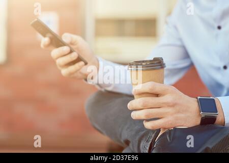 Zugeschnittene Aufnahme eines Geschäftsmannes in blauem Hemd mit Smartphone Und Kaffee trinken, während Sie sich auf der Bank im Freien entspannen Stockfoto