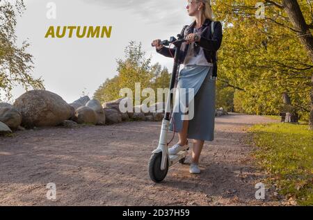 Eine junge Blondine steht im Frühherbst mit einem Elektroroller im Park. Stockfoto