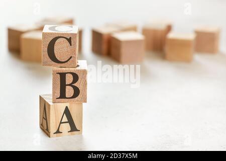 Drei Holzwürfel mit den Buchstaben CBA (bedeutet Kosten-Nutzen-Bewertung), auf weißem Tisch, mehr im Hintergrund, Platz für Text in der rechten unteren Ecke Stockfoto