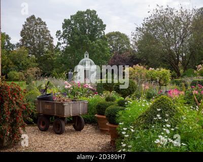 Chenies Manor Garten. Ein Handwagen ruht nach der Arbeit im Garten auf dem Weg. Ein Vogelkäfigpavillon im Parterre hinter den rosa und weißen Blüten. Stockfoto
