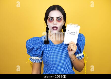 Frau trägt ein gruseliges Puppe halloween Kostüm auf gelbem Hintergrund Erschrocken und überrascht halten Notebook ohne Wort Nachricht Stockfoto