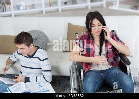 Junge Frau im Rollstuhl auf Smartphone junger Mann Hausaufgaben machen Stockfoto