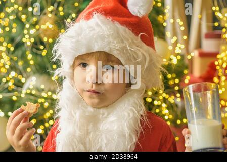 Kid Santa Claus genießen in serviert Lebkuchen Kuchen und Milch. Kid Santa Claus nimmt ein Cookie am Heiligabend als Dankeschön Geschenk für das Verlassen Geschenke Stockfoto