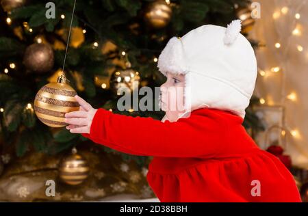 Das Baby greift nach einem Weihnachtsbaumball. Ein nettes kleines Mädchen in einem roten Kleid und weißem Hut drückt Emotionen aus. Weihnachtskonzept mit kleinem Kind, tre Stockfoto