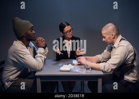 Ein Drogenhändler, der mit einem Paket mit einer unbekannten weißen Substanz inhaftiert ist, wird von einem erfahrenen Ermittler in Anwesenheit eines Rechtsanwalts verhört. Stockfoto