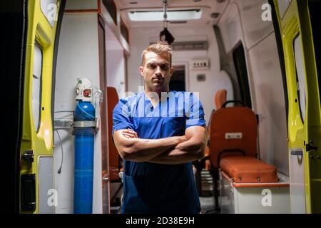 Mann in einer medizinischen Uniform, der ernsthaft auf die Kamera schaut, Arme gefaltet, Ambulanzwagen im Hintergrund. Stockfoto