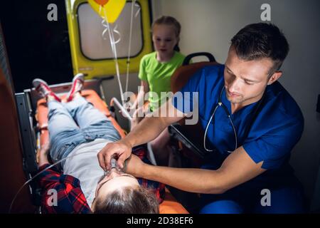 Junger Mann in einer medizinischen Uniform, der eine Sauerstoffmaske auf dem Gesicht einer Frau hält, die auf einer Bahre liegt, kleines trauriges Mädchen, das neben ihr in einem Krankenwagen sitzt. Stockfoto